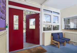 Image of double red doors with one open inside a room with blue carpet and chairs