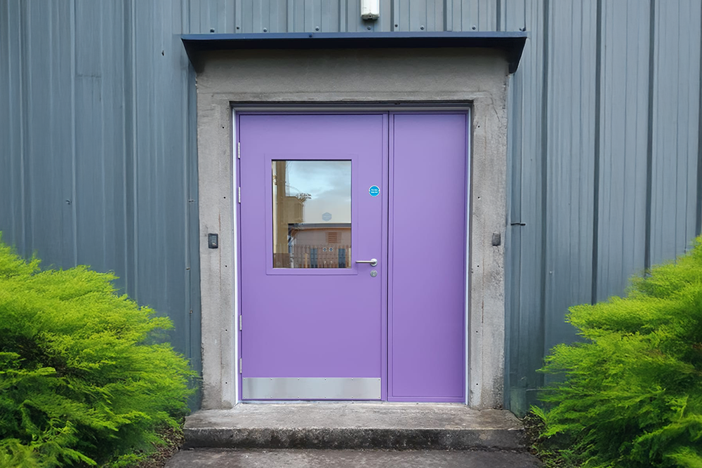 A purple fire rated external door against a grey wall with bushes on either side.